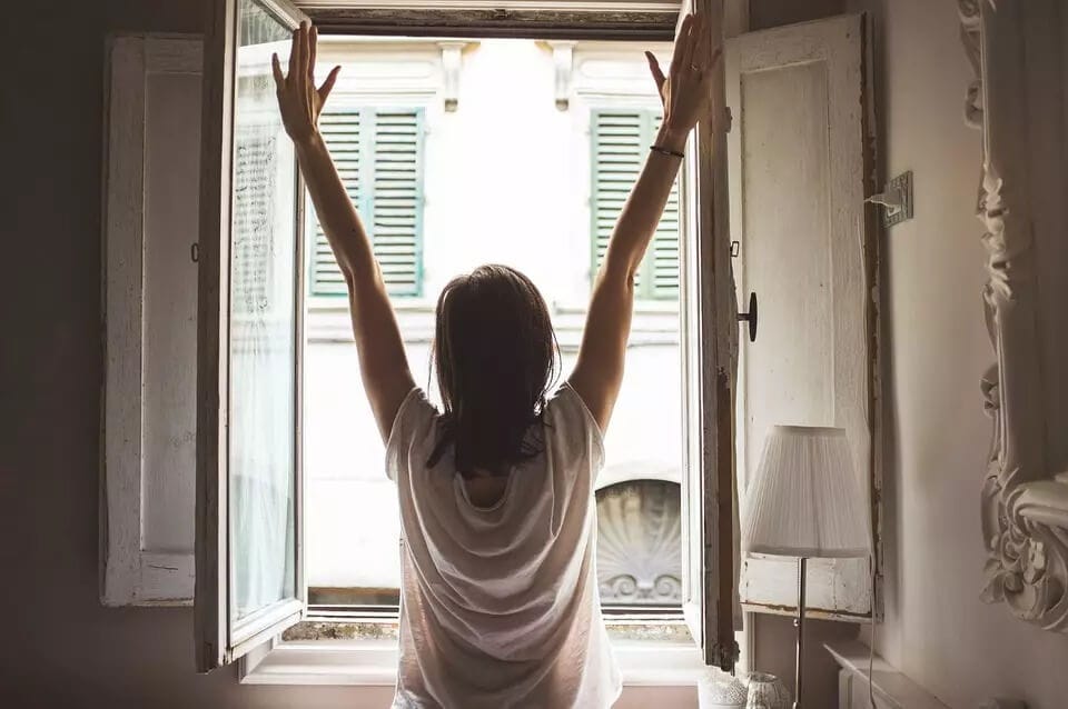 Woman facing window stretching with arms in the air